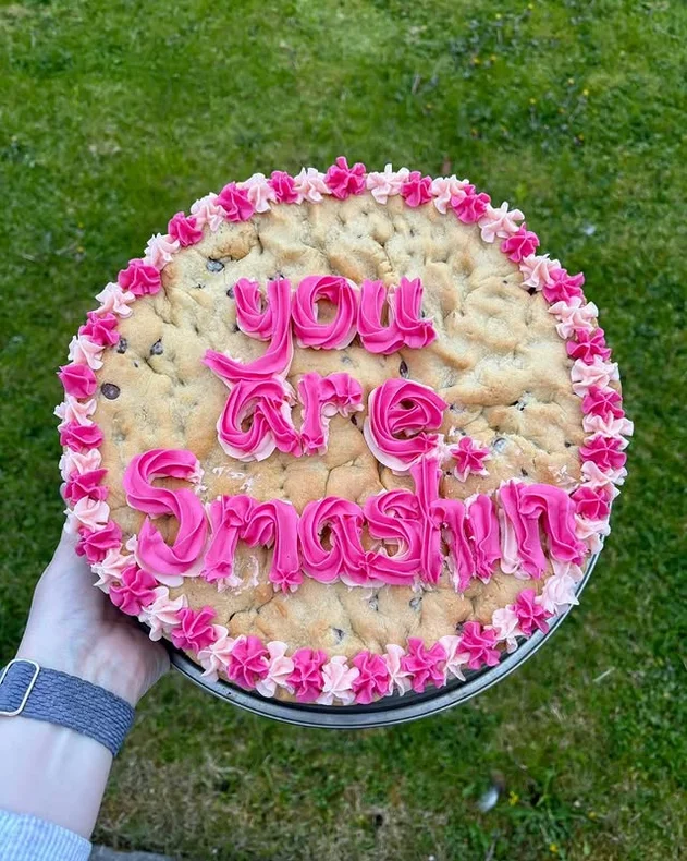 A giant cookie cake with pink piped lettering and a pink and white border